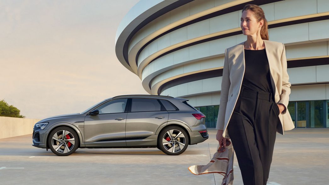 Side view of a parked, grey Audi Q8 e-tron with a building in the background and woman in the front
