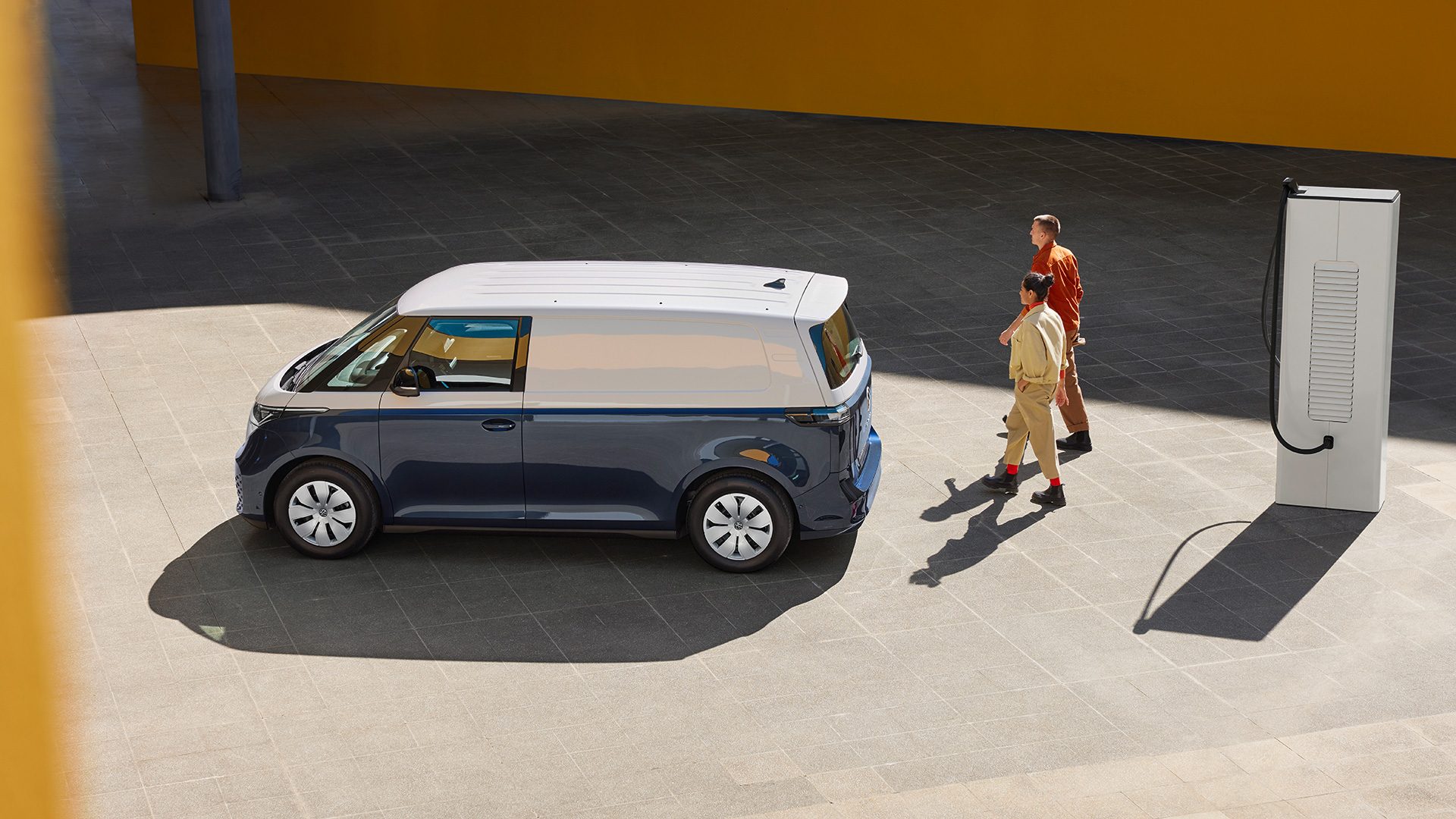AMAG VW ID. Buzz in bianco blu, vista laterale dall'alto, due persone che camminano tra l'auto e la stazione di ricarica